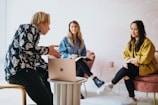 man in black and white floral long sleeve shirt sitting and showing something on a microsoft laptop to 2 women 