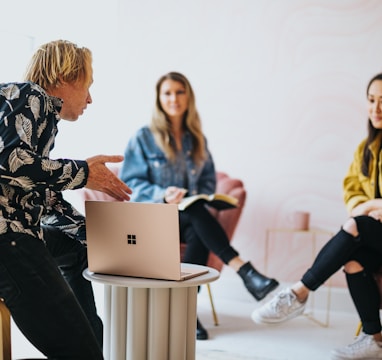 man in black and white floral long sleeve shirt sitting and showing something on a microsoft laptop to 2 women 