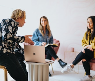 man in black and white floral long sleeve shirt sitting and showing something on a microsoft laptop to 2 women 