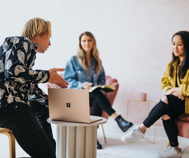 man in black and white floral long sleeve shirt sitting and showing something on a microsoft laptop to 2 women 