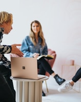 man in black and white floral long sleeve shirt sitting and showing something on a microsoft laptop to 2 women 