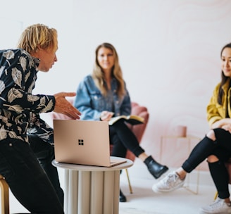 man in black and white floral long sleeve shirt sitting and showing something on a microsoft laptop to 2 women 