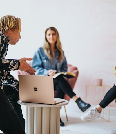 man in black and white floral long sleeve shirt sitting and showing something on a microsoft laptop to 2 women 