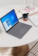 A sleek laptop with a grey keyboard rests on a round white table. The screen displays the Windows operating system interface. Nearby, there is a ceramic mug with a colorful pattern, a notebook with handwritten notes, and an open book with floral illustrations. The surface of the table is smooth and clean, with a warm light reflecting off it.
