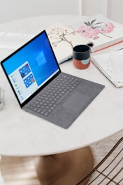 A sleek laptop with a grey keyboard rests on a round white table. The screen displays the Windows operating system interface. Nearby, there is a ceramic mug with a colorful pattern, a notebook with handwritten notes, and an open book with floral illustrations. The surface of the table is smooth and clean, with a warm light reflecting off it.