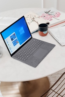 A sleek laptop with a grey keyboard rests on a round white table. The screen displays the Windows operating system interface. Nearby, there is a ceramic mug with a colorful pattern, a notebook with handwritten notes, and an open book with floral illustrations. The surface of the table is smooth and clean, with a warm light reflecting off it.