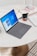A sleek laptop with a grey keyboard rests on a round white table. The screen displays the Windows operating system interface. Nearby, there is a ceramic mug with a colorful pattern, a notebook with handwritten notes, and an open book with floral illustrations. The surface of the table is smooth and clean, with a warm light reflecting off it.