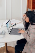 A person with long hair and glasses is seated at a round table, focused on a laptop displaying graphs. Open books and a pair of headphones are also on the table, suggesting a study or work setting.