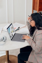 A person with long hair and glasses is seated at a round table, focused on a laptop displaying graphs. Open books and a pair of headphones are also on the table, suggesting a study or work setting.