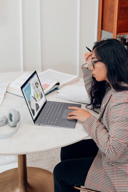 A person with long hair and glasses is seated at a round table, focused on a laptop displaying graphs. Open books and a pair of headphones are also on the table, suggesting a study or work setting.