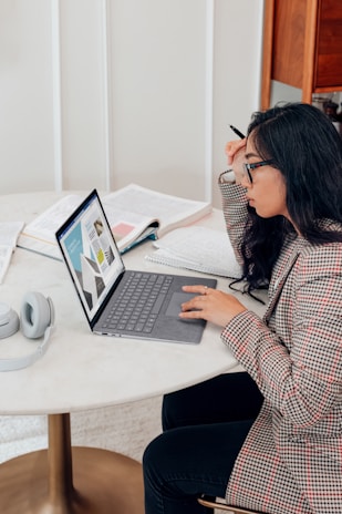 A person with long hair and glasses is seated at a round table, focused on a laptop displaying graphs. Open books and a pair of headphones are also on the table, suggesting a study or work setting.