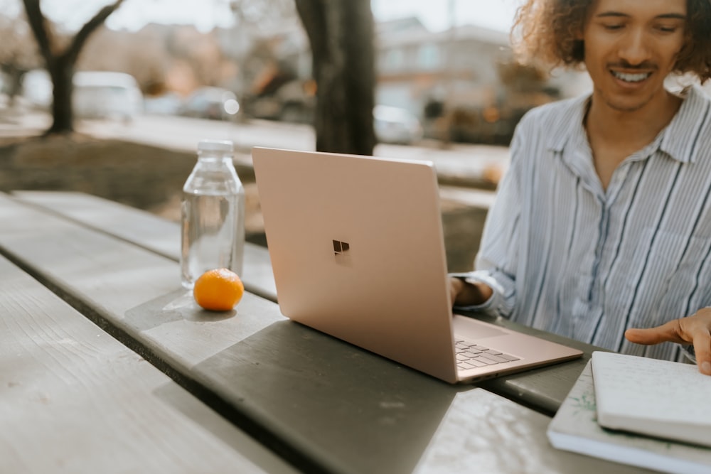 昼間、緑の木製のテーブルで Microsoft Surface ノート PC を使用する青いドレス シャツを着た男性