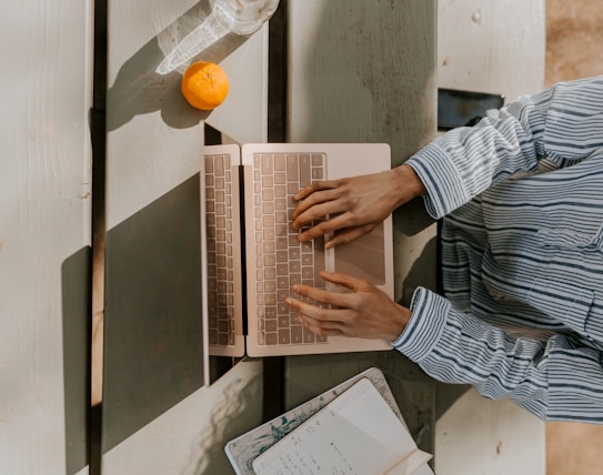 A person in a striped shirt is typing on a rose-gold laptop placed on a wooden picnic table. There's a notebook with writing and a small orange fruit next to the laptop. Sunlight casts shadows across the table, creating a tranquil and focused atmosphere.