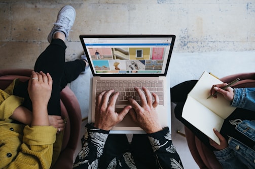 Three people sitting together, one using a laptop on their lap and the others taking notes. Images displayed on the laptop screen feature various scenes, including a bicycle and colorful buildings. The floor is light, and the individuals have casual attire.