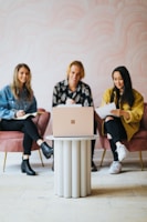 Three people are seated on pink chairs around a small, modern circular table with a laptop placed on top. They appear to be engaged, taking notes and smiling, suggesting a collaborative or meeting environment.