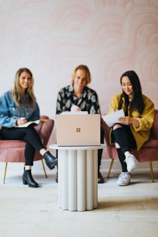 Three people are seated on pink chairs around a small, modern circular table with a laptop placed on top. They appear to be engaged, taking notes and smiling, suggesting a collaborative or meeting environment.