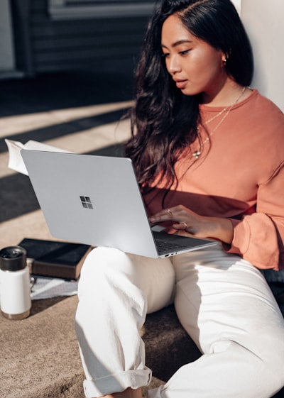 woman in orange long sleeve shirt and white pants sitting on floor using Surface Laptop 