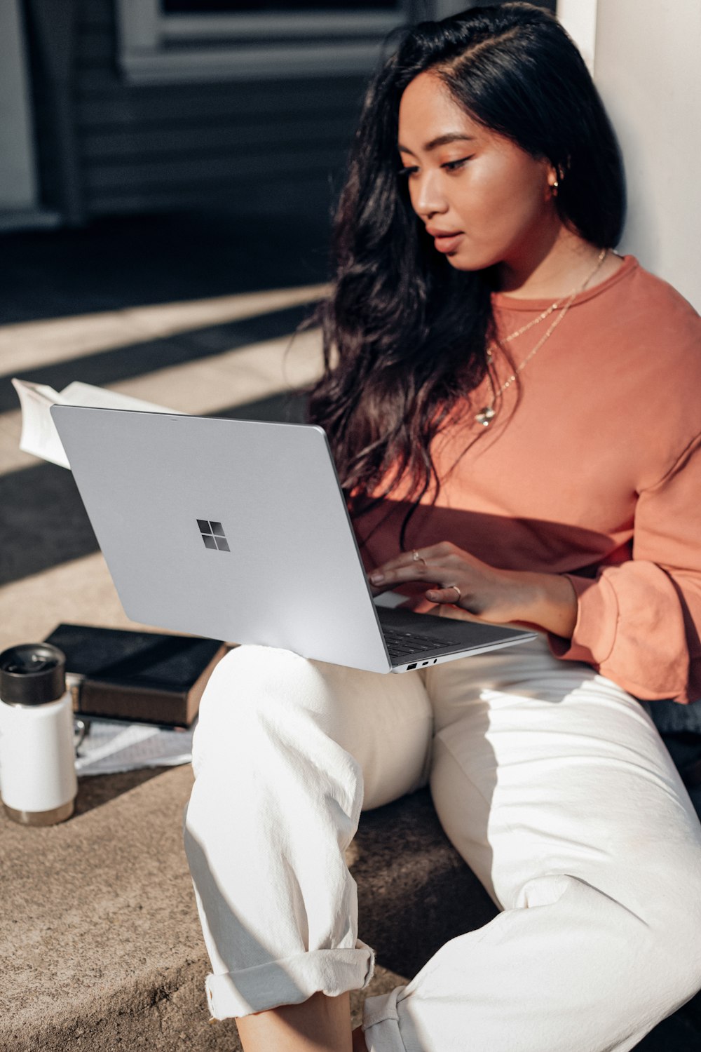 オレンジ色の長袖シャツと白いズボンを着た女性が床に座り、Surface Laptop を使用している