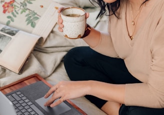 woman in beige long sleeve shirt and black pants sitting on bed using a Microsoft Surface Laptop 3 