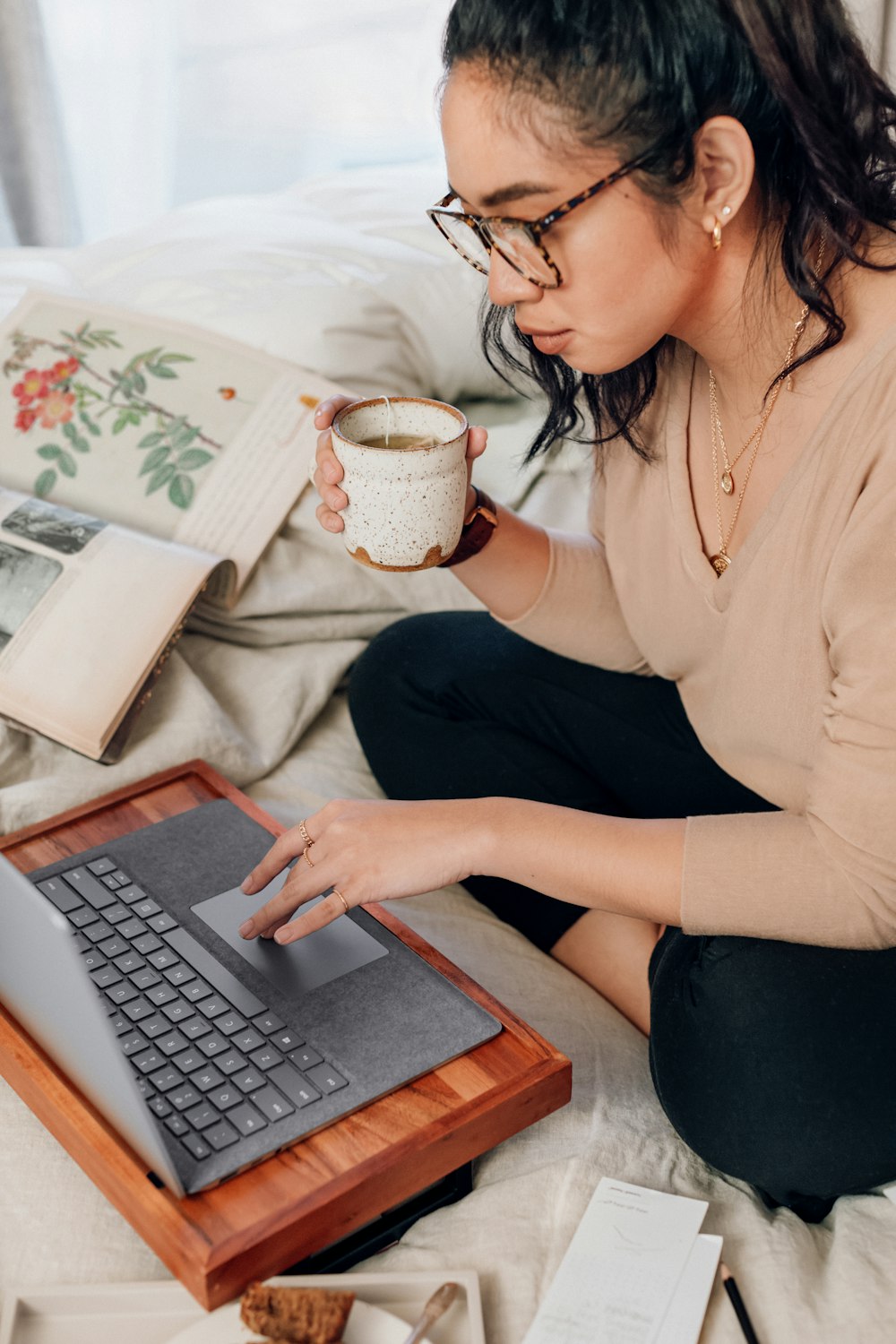 ベージュの長袖シャツと黒いズボンを着た女性がベッドに座って Microsoft Surface Laptop 3 を使用している