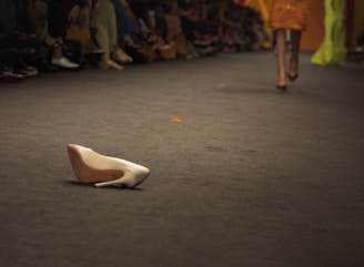 A high-heeled white shoe is lying on a runway with blurred fashion models walking in the background. Spectators are seated on either side of the runway.