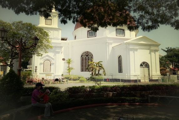 A white, historic-style church with large arched windows and a domed roof is surrounded by a small, well-kept garden. The church is fenced, and there are lush green trees providing shade in the foreground. A person is sitting on a bench with a child, creating a peaceful and family-friendly atmosphere.