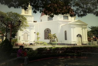 A white, historic-style church with large arched windows and a domed roof is surrounded by a small, well-kept garden. The church is fenced, and there are lush green trees providing shade in the foreground. A person is sitting on a bench with a child, creating a peaceful and family-friendly atmosphere.