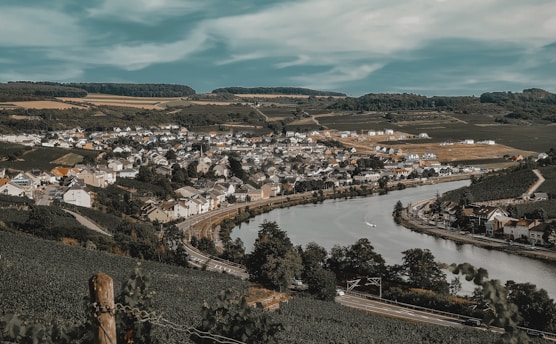 A picturesque village nestled within a lush, green valley bordered by vineyards. The village is situated along a winding river, with neatly arranged houses and buildings. Rolling hills and forests surround the area, creating a serene landscape under a partly cloudy sky.