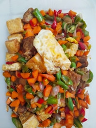 A colorful mix of cut vegetables and tofu is presented on a plate, featuring bright orange carrots, green beans, and red peppers. There is a piece of fried egg on top, adding a contrasting texture and color.