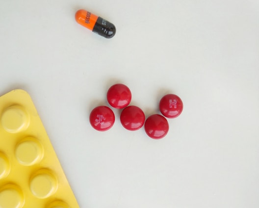 Five red round pills are arranged in a circular pattern on a light surface. Nearby, there is a bicolored orange and black capsule with the word 'BECOBA' written on it. A yellow blister pack with circular indentations is partially visible in the bottom left corner.
