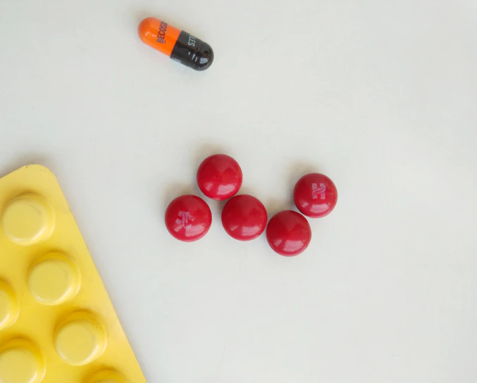 Five red round pills are arranged in a circular pattern on a light surface. Nearby, there is a bicolored orange and black capsule with the word 'BECOBA' written on it. A yellow blister pack with circular indentations is partially visible in the bottom left corner.