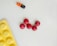 Five red round pills are arranged in a circular pattern on a light surface. Nearby, there is a bicolored orange and black capsule with the word 'BECOBA' written on it. A yellow blister pack with circular indentations is partially visible in the bottom left corner.