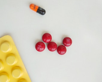 Five red round pills are arranged in a circular pattern on a light surface. Nearby, there is a bicolored orange and black capsule with the word 'BECOBA' written on it. A yellow blister pack with circular indentations is partially visible in the bottom left corner.