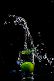A lime is submerged in a small glass of water, creating splashes that rise above it. The background is completely black, highlighting the vivid green of the lime and the clear water droplets.