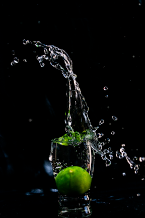 A lime is submerged in a small glass of water, creating splashes that rise above it. The background is completely black, highlighting the vivid green of the lime and the clear water droplets.