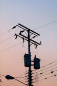 A utility pole with multiple wires and cylindrical transformers is silhouetted against a gradient sky. The surrounding cables are slightly tangled, and a street lamp is visible at the bottom left corner. The overall scene suggests early morning or late evening due to the soft pastel colors in the sky.