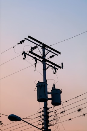 A utility pole with multiple wires and cylindrical transformers is silhouetted against a gradient sky. The surrounding cables are slightly tangled, and a street lamp is visible at the bottom left corner. The overall scene suggests early morning or late evening due to the soft pastel colors in the sky.