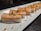 A platter of neatly arranged sushi pieces featuring a layer of seared salmon on top of rice, garnished with sauce and a small crispy topping. The sushi pieces are placed in a row on a rectangular white plate with a dark background.