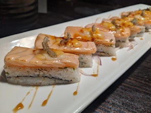 A platter of neatly arranged sushi pieces featuring a layer of seared salmon on top of rice, garnished with sauce and a small crispy topping. The sushi pieces are placed in a row on a rectangular white plate with a dark background.