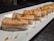 A platter of neatly arranged sushi pieces featuring a layer of seared salmon on top of rice, garnished with sauce and a small crispy topping. The sushi pieces are placed in a row on a rectangular white plate with a dark background.
