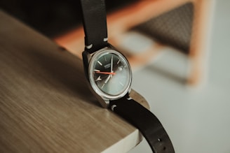A classic wristwatch with a black leather strap rests on the corner of a wooden surface. The watch face is round with dark tones and metallic accents, featuring a red second hand.