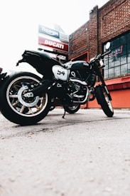 A motorcycle is parked outdoors in front of a building with signs for Triumph and Ducati. The motorcycle is black with the number 54 displayed on its side, and it features a retro design with gold-colored rims. The setting appears to be an urban area with a brick building in the background and large windows.
