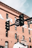 A traffic light and street signs set against the backdrop of a historic red brick building. The building has several windows and faded signage. The street sign mentions 'Denver' and another sign reads 'Do Not Enter' with exceptions for buses.