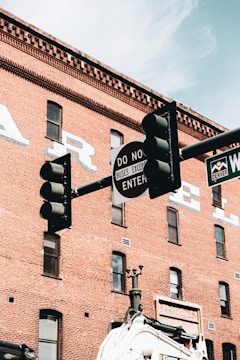 A traffic light and street signs set against the backdrop of a historic red brick building. The building has several windows and faded signage. The street sign mentions 'Denver' and another sign reads 'Do Not Enter' with exceptions for buses.