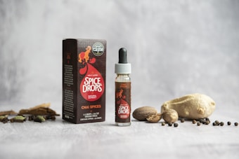 A small bottle labeled 'Spice Drops' placed next to a cardboard box with the same branding, surrounded by an assortment of whole spices, including ginger, nutmeg, and peppercorns, on a textured light gray surface.