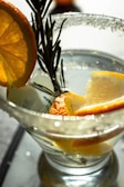 A clear cocktail glass contains a light-colored liquid, garnished with slices of orange and a sprig of rosemary. Sugar rims the edge of the glass, which captures the light creating soft reflections. The oranges add a burst of color and freshness to the sophisticated presentation.