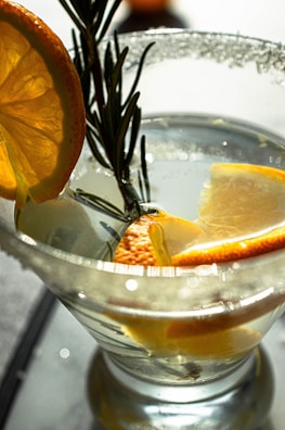 A clear cocktail glass contains a light-colored liquid, garnished with slices of orange and a sprig of rosemary. Sugar rims the edge of the glass, which captures the light creating soft reflections. The oranges add a burst of color and freshness to the sophisticated presentation.