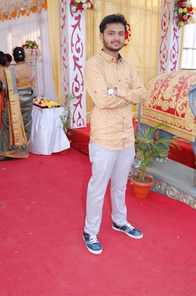 A young man stands with arms crossed on a red carpet, wearing a beige patterned shirt and gray trousers with blue sneakers. The setting is festive, featuring ornate pillars decorated with red and white patterns, and floral arrangements. A woman in a sari is visible in the background, along with a decorative seat or palanquin.
