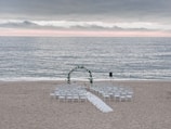 A wedding ceremony setup on a beach features an archway adorned with greenery and white flowers, surrounded by rows of white chairs arranged symmetrically facing the arch. The ocean waves gently roll in the background under a cloudy sky with a hint of sunlight breaking through the clouds along the horizon.