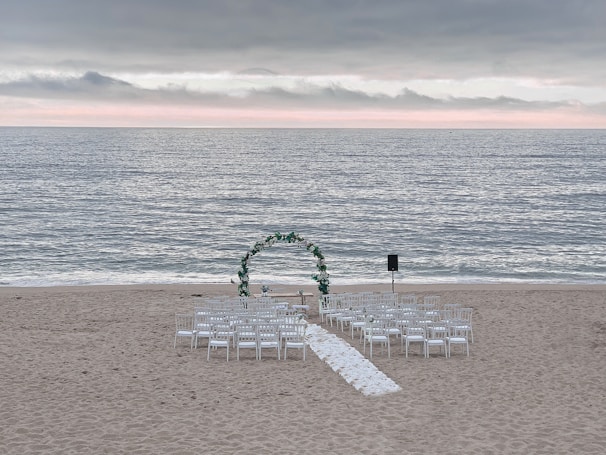 A wedding ceremony setup on a beach features an archway adorned with greenery and white flowers, surrounded by rows of white chairs arranged symmetrically facing the arch. The ocean waves gently roll in the background under a cloudy sky with a hint of sunlight breaking through the clouds along the horizon.